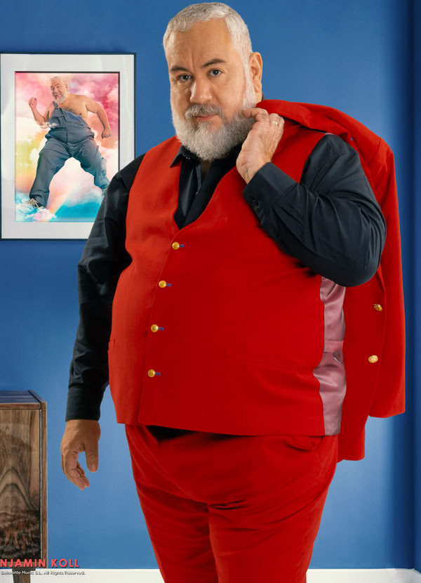 Man with a beard wearing a red suit and holding the jacket over his shoulder, standing in a room with blue walls and a framed picture on the wall.