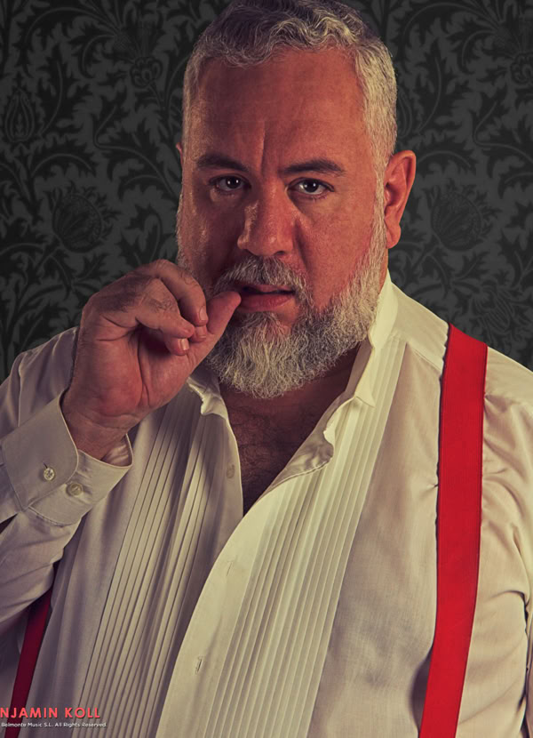 A man with a beard and gray hair wearing a white shirt and red suspenders, standing against a dark patterned background, touching his lips with his finger.
