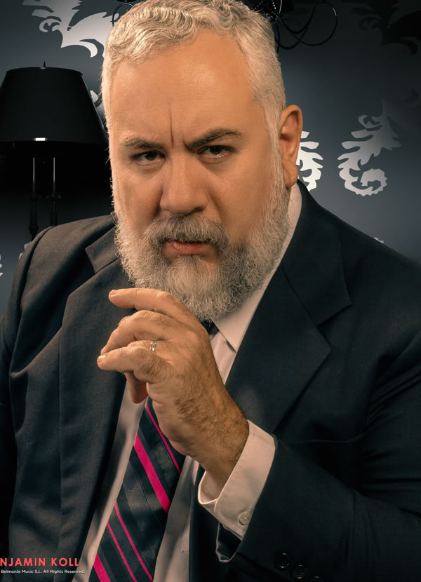 A man with a gray beard wearing a suit and tie, sitting in front of a patterned wallpaper with a lamp in the background.
