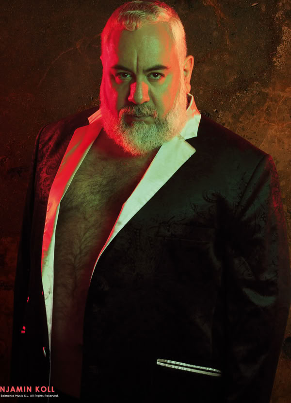 A man with a beard wearing a black suit jacket with white lapels, standing against a textured background with red and green lighting.