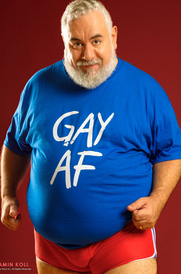 A person with a beard wearing a blue shirt with "GAY AF" written on it and red shorts, standing against a red background.