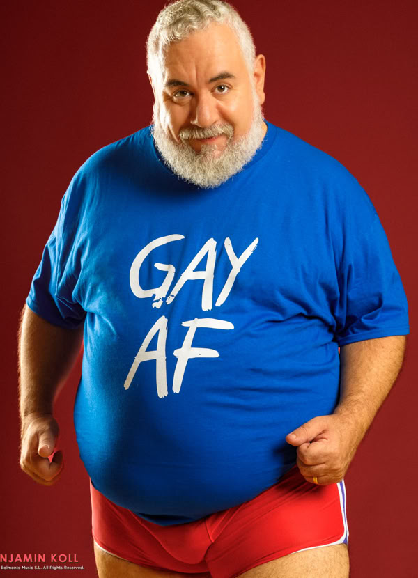A person with a beard wearing a blue shirt with "GAY AF" written on it and red shorts, standing against a red background.