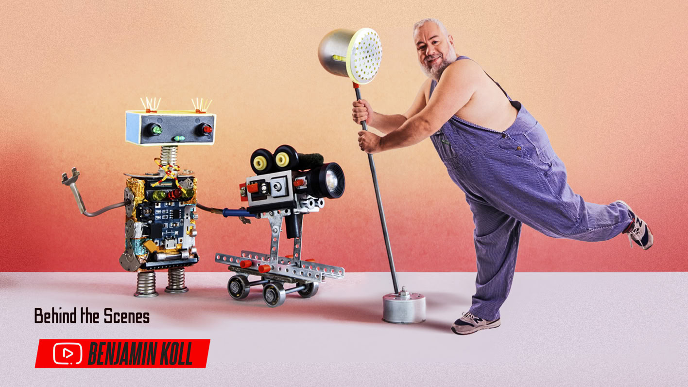 A person in overalls holds a large mallet, standing next to two robots; one with a camera and the other with a screen for a head, against a gradient background.