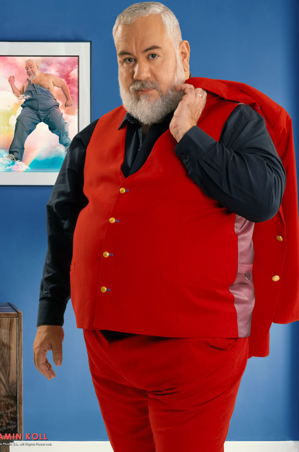 Man with a beard wearing a red suit and holding the jacket over his shoulder, standing in a room with blue walls and a framed picture on the wall.