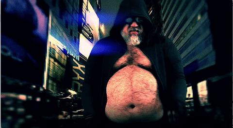 A person wearing a hooded jacket stands in an urban setting at night, with tall buildings and bright lights in the background.